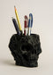 Black skull-shaped pen holder with various pens on a light beige background